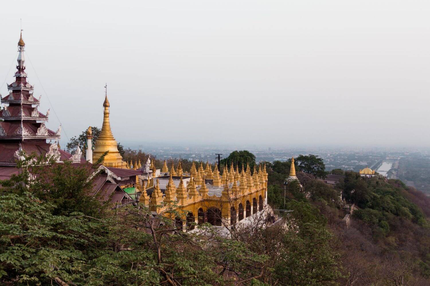 Best Myanmar Itinerary: Photo of the Mandalay Hill and Temple in Mandalay Myanmar, with the golden spires pointing over the trees atop the mountain overlooking Mandalay. Photo taken by Ryan Brown of Lost Boy Memoirs, edited in Lightroom.