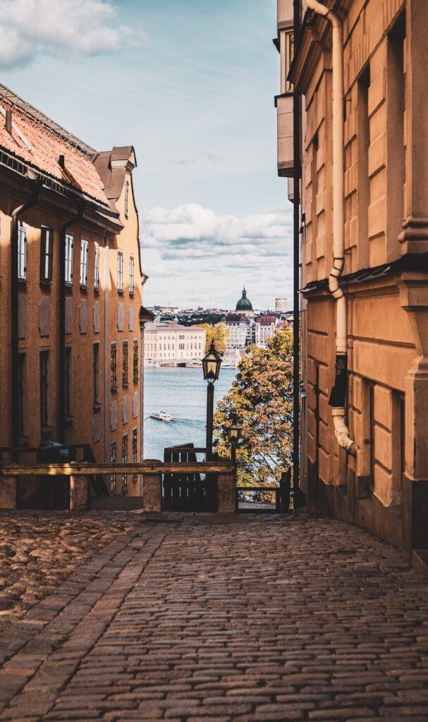 A view of a cobblestone street in Stockholm at sunset between two buildings with a view of the sea in the distance.