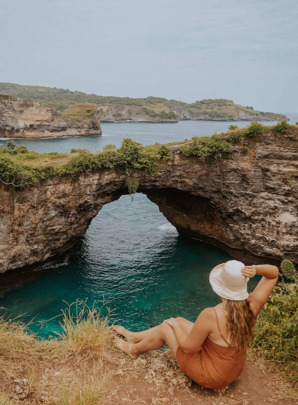 A woman sitting on the edge of a cliff wearing a white straw hat overlooking the famous Broken Beach on Nusa Penida island in Bali
