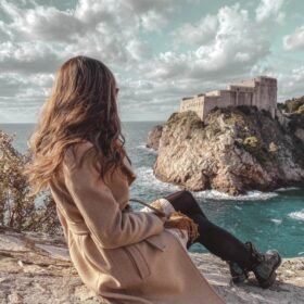 A woman sitting on the walls of Dubrovnik looking out at the Lovrijenac Fortress and Adriatic Sea.